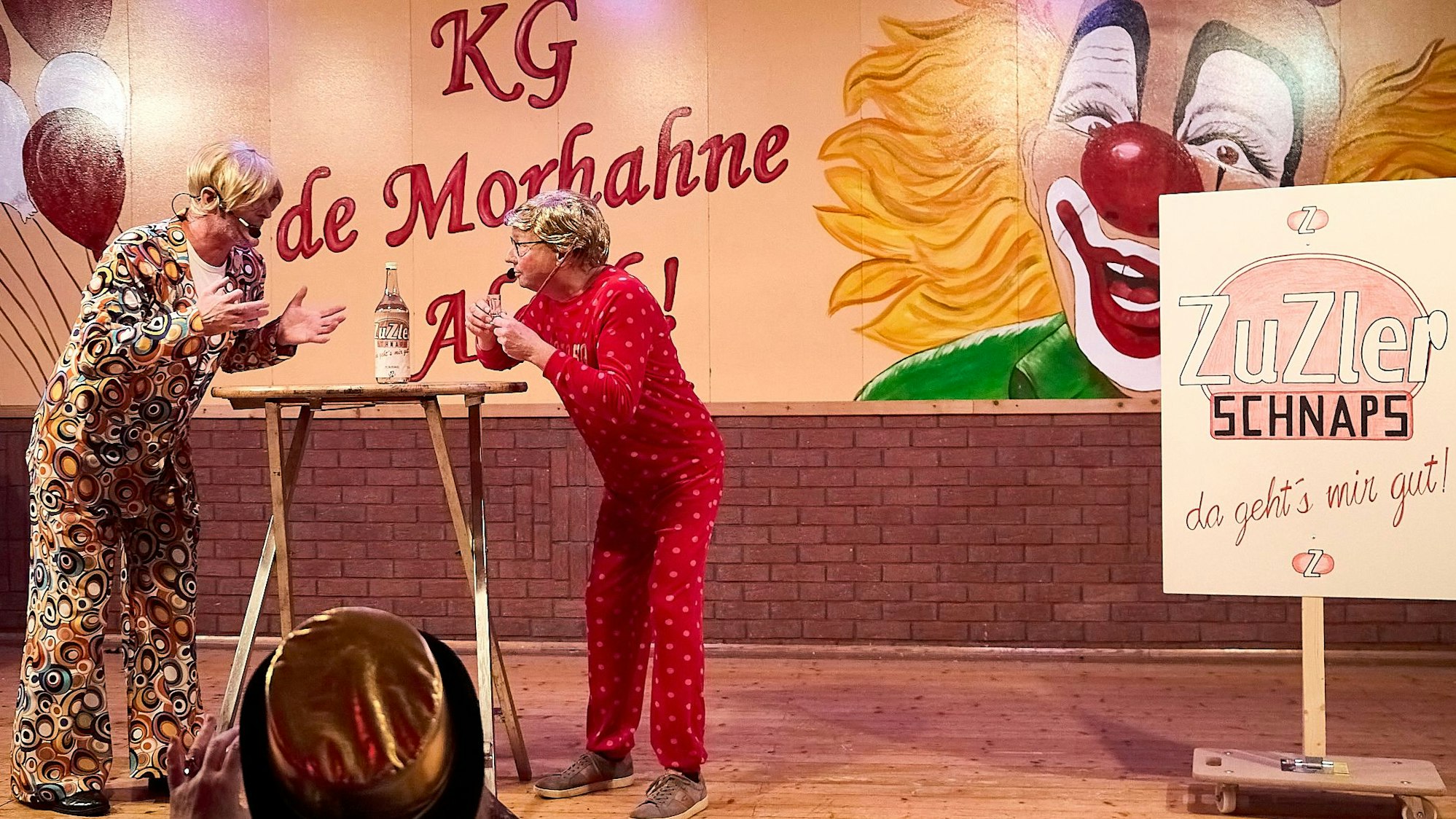 Ralf Meyer und Norbert Becker stehen in bunten Kostümen an einem Tisch, auf dem sich eine Schnapsflasche befindet.
