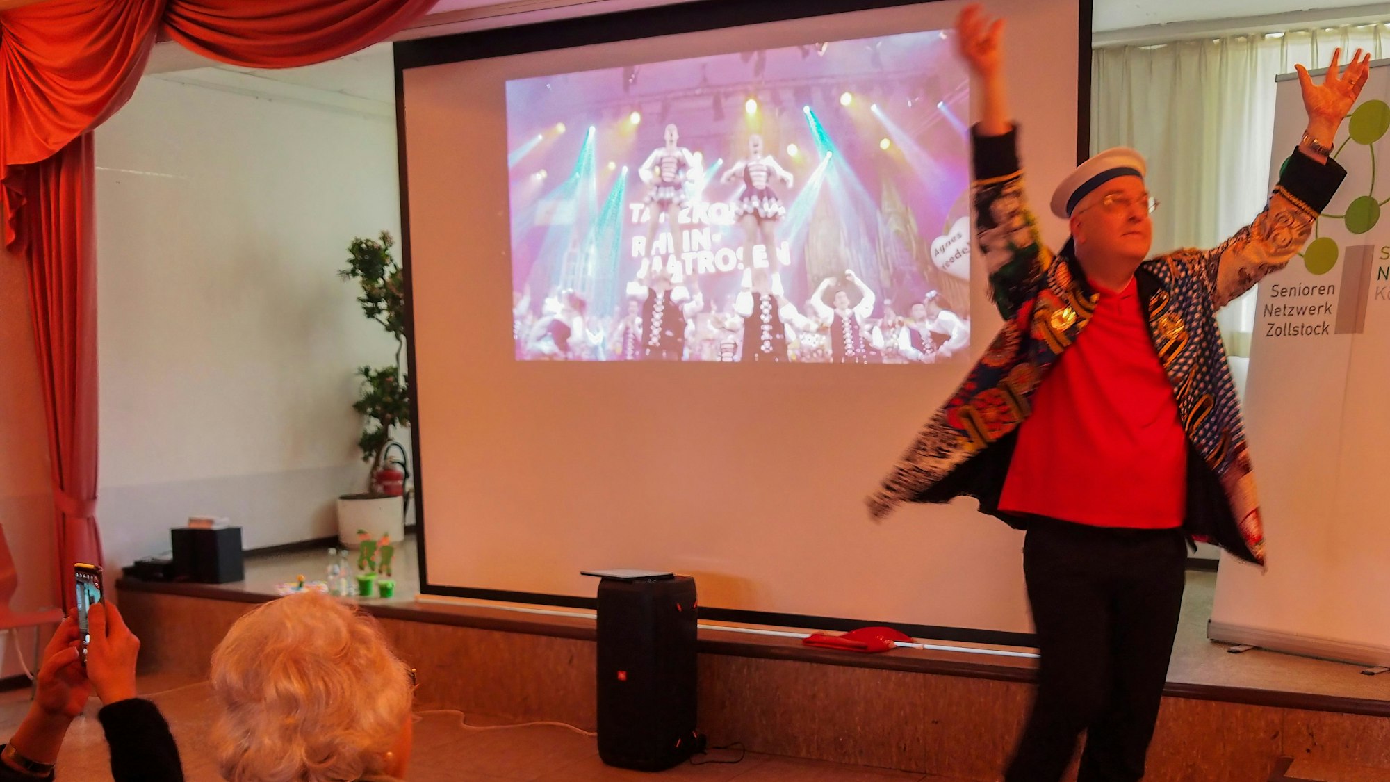 Uli Kievernagel steht mit erhobenen Armen auf der Bühne, hinter ihm auf einer Leinwand ist ein Tanzcorps zu sehen.