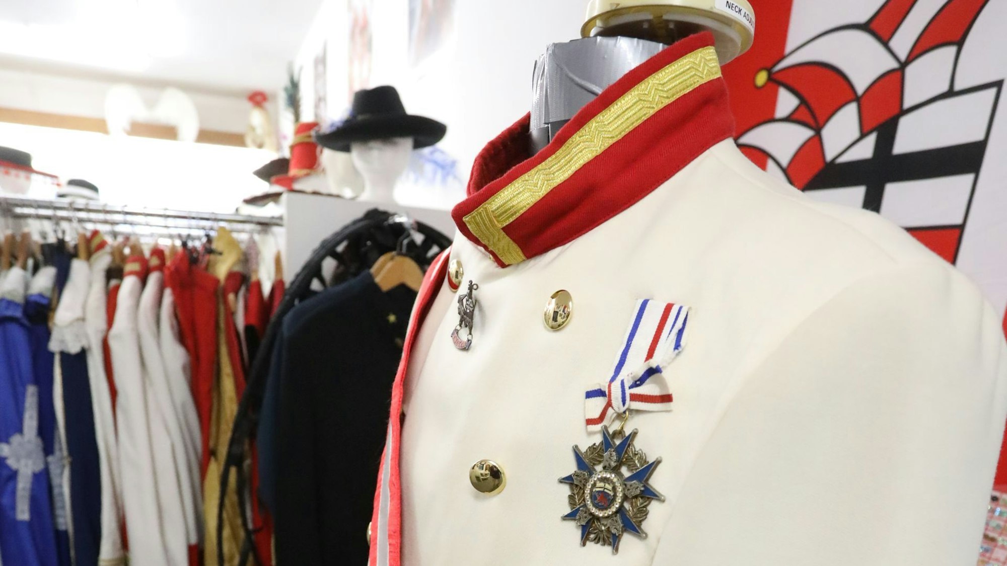 Eine weiße Uniformjacke mit großem Orden, im Hintergrund eine Schaufensterpuppe und Kostüme auf einem Ständer.