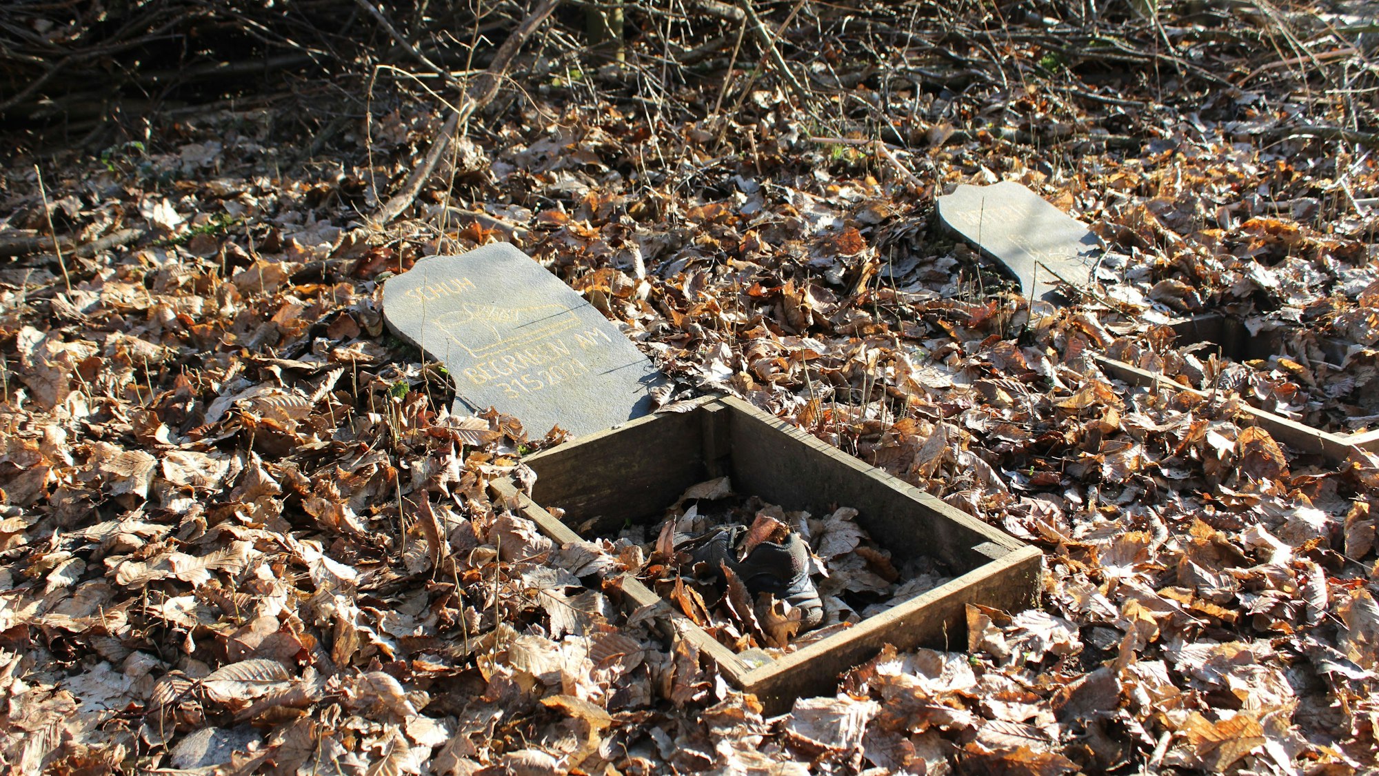 Umgeben von Laub liegt ein Schuh in einem Holzkasten, der mit Laub und Erde gefüllt ist. Auf einem Grabstein darüber steht „Schuh: Begraben am 31.5.2021“.