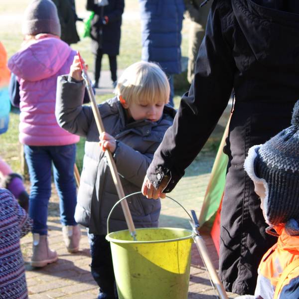 Zwei Kinder legt mit ihren Müllzangen etwas in einen Eimer, den ihnen eine Erzieherin hinhält, im Hintergrund sind weitere Kinder zu sehen, die eine Wiese nach Müll absuchen.