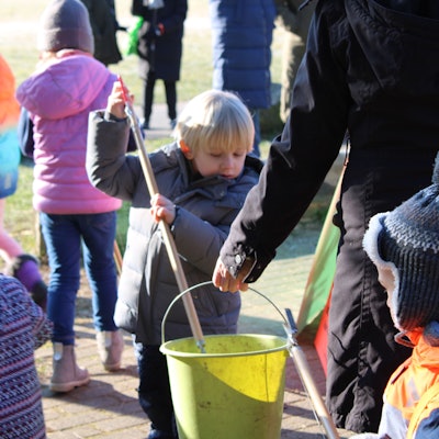 Zwei Kinder legt mit ihren Müllzangen etwas in einen Eimer, den ihnen eine Erzieherin hinhält, im Hintergrund sind weitere Kinder zu sehen, die eine Wiese nach Müll absuchen.