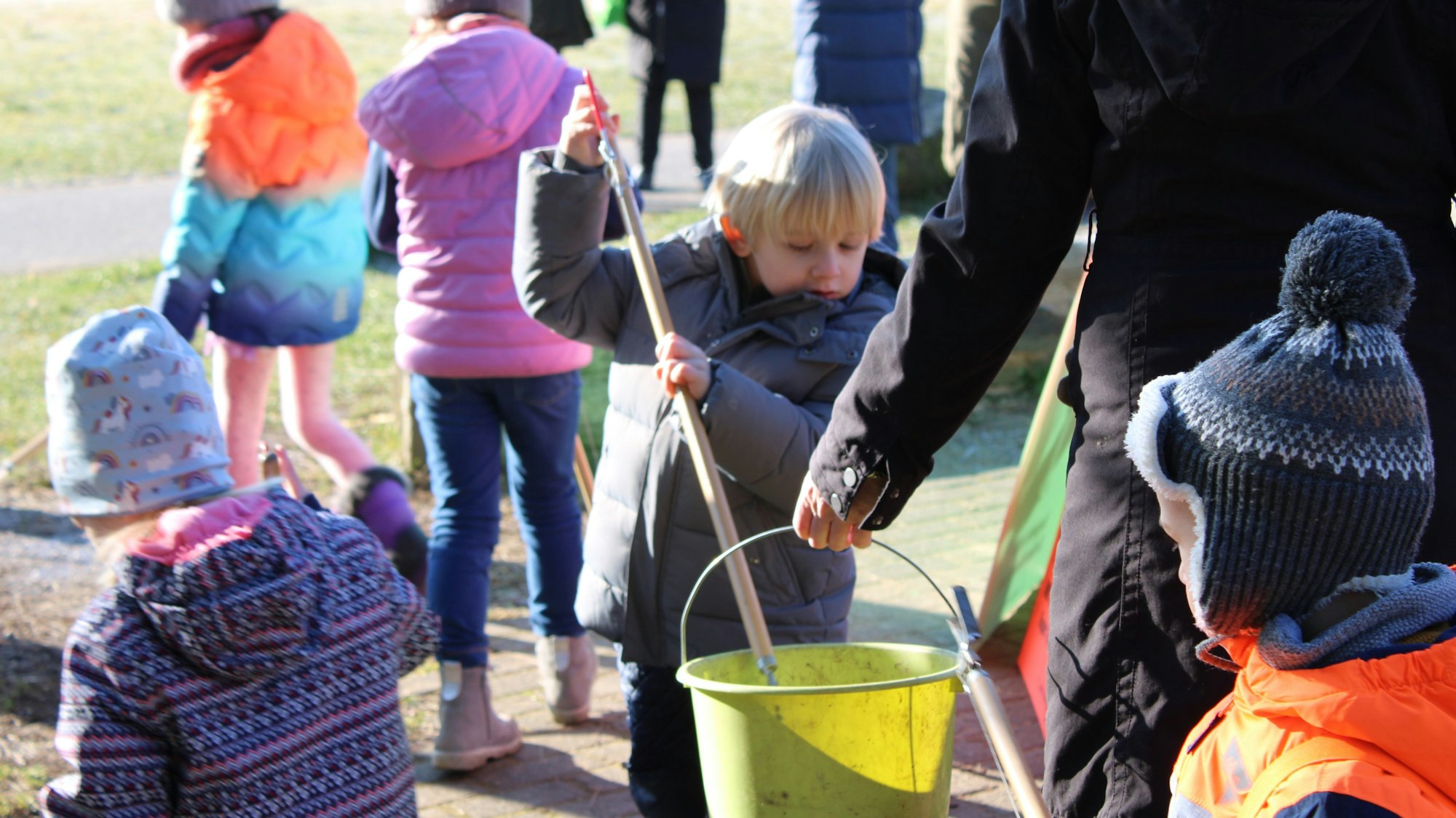 Zwei Kinder legt mit ihren Müllzangen etwas in einen Eimer, den ihnen eine Erzieherin hinhält, im Hintergrund sind weitere Kinder zu sehen, die eine Wiese nach Müll absuchen.