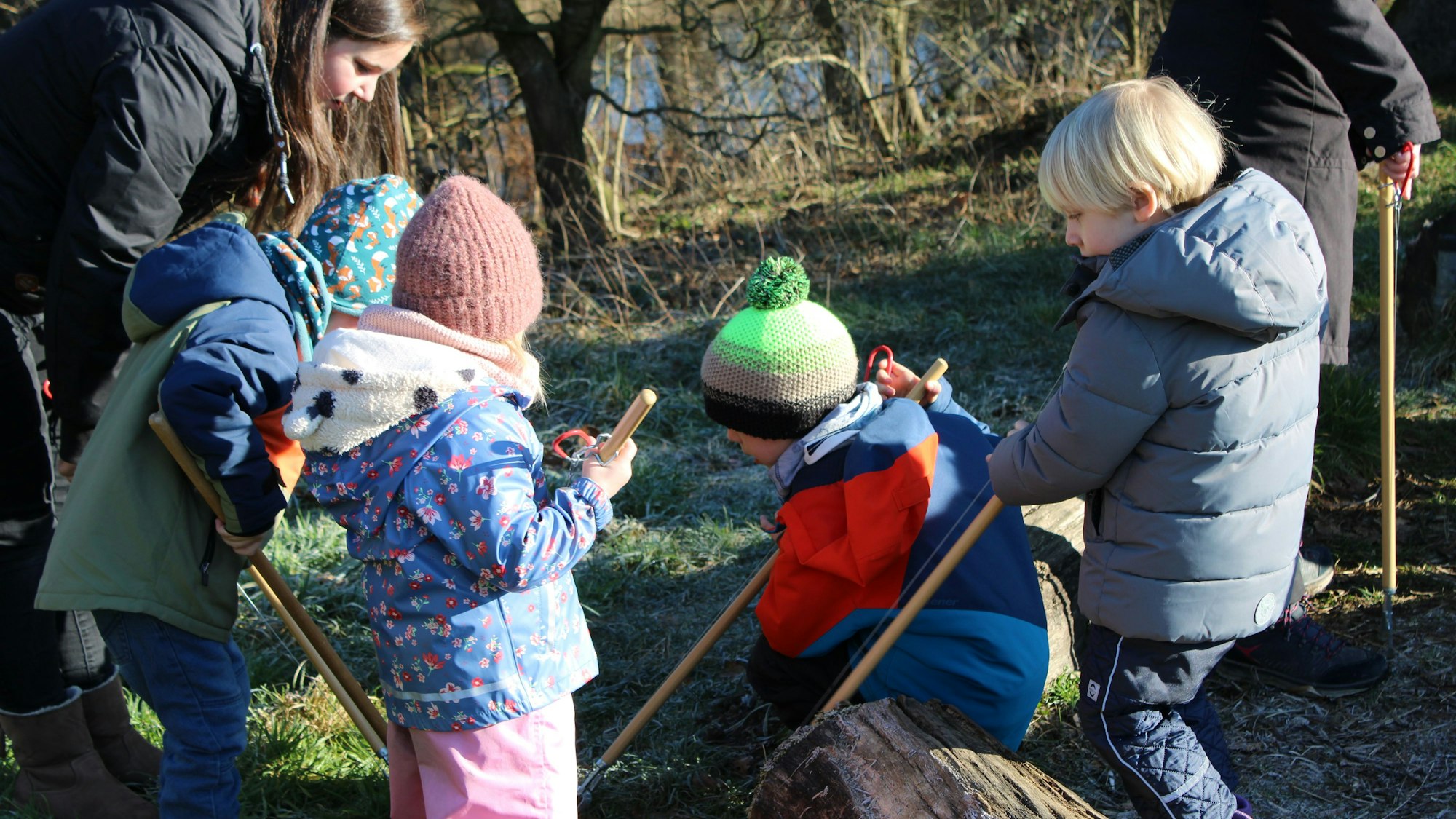 Vier Kinder suchen mit Müllzangen eine Wiese ab, eine Erzieherin steht neben ihnen.