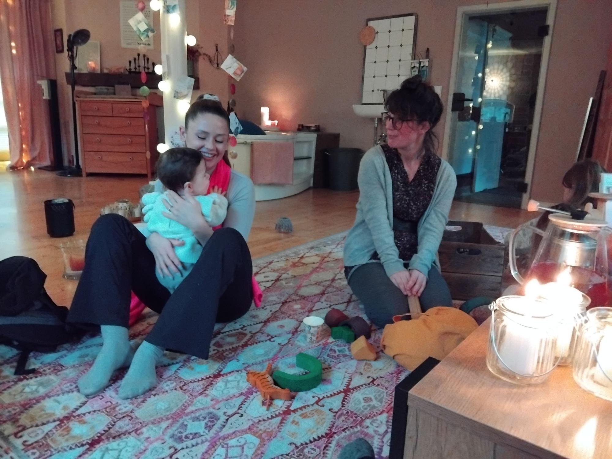 Eine Frau mit Baby sitzt auf einem Teppich, eine zweite kniet neben ihr, im Hintergrund steht eine große Badewanne