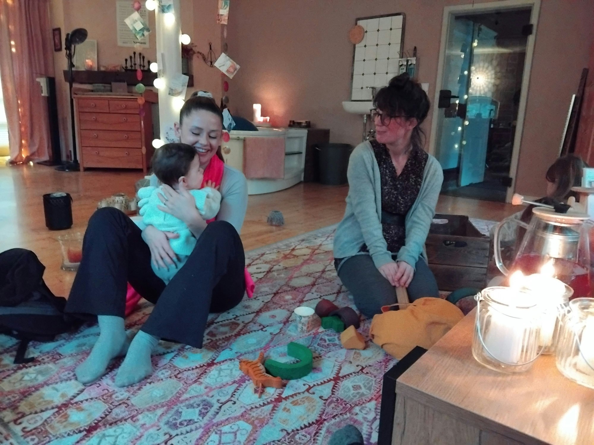 Eine Frau mit Baby sitzt auf einem Teppich, eine zweite kniet neben ihr, im Hintergrund steht eine große Badewanne
