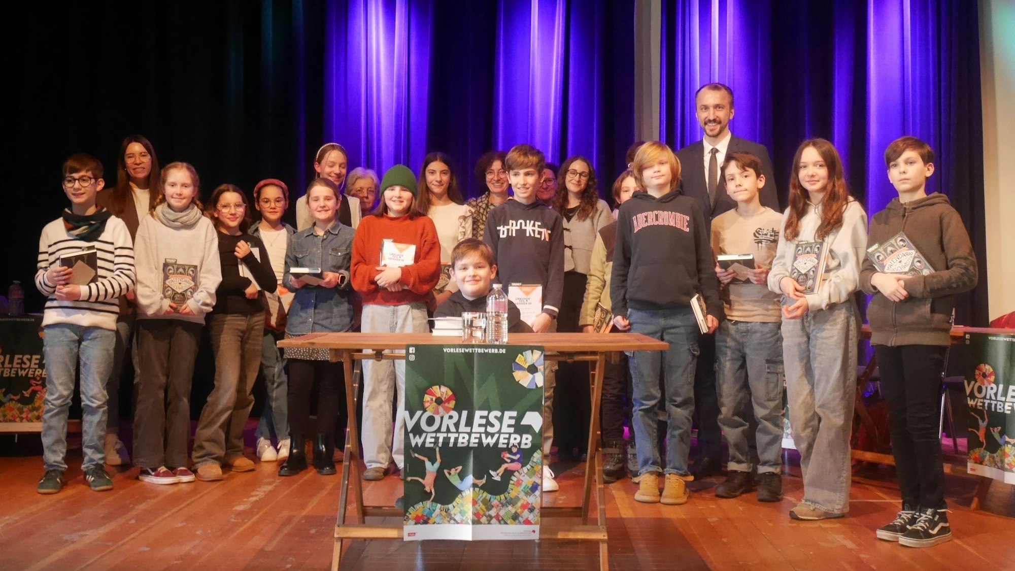 Die Kinder stehen auf der Bühne, der Gewinner sitzt an einem Tisch vor ihnen. Ein Plakat am Tisch trägt die Aufschrift „Vorlesewettbewerb“.