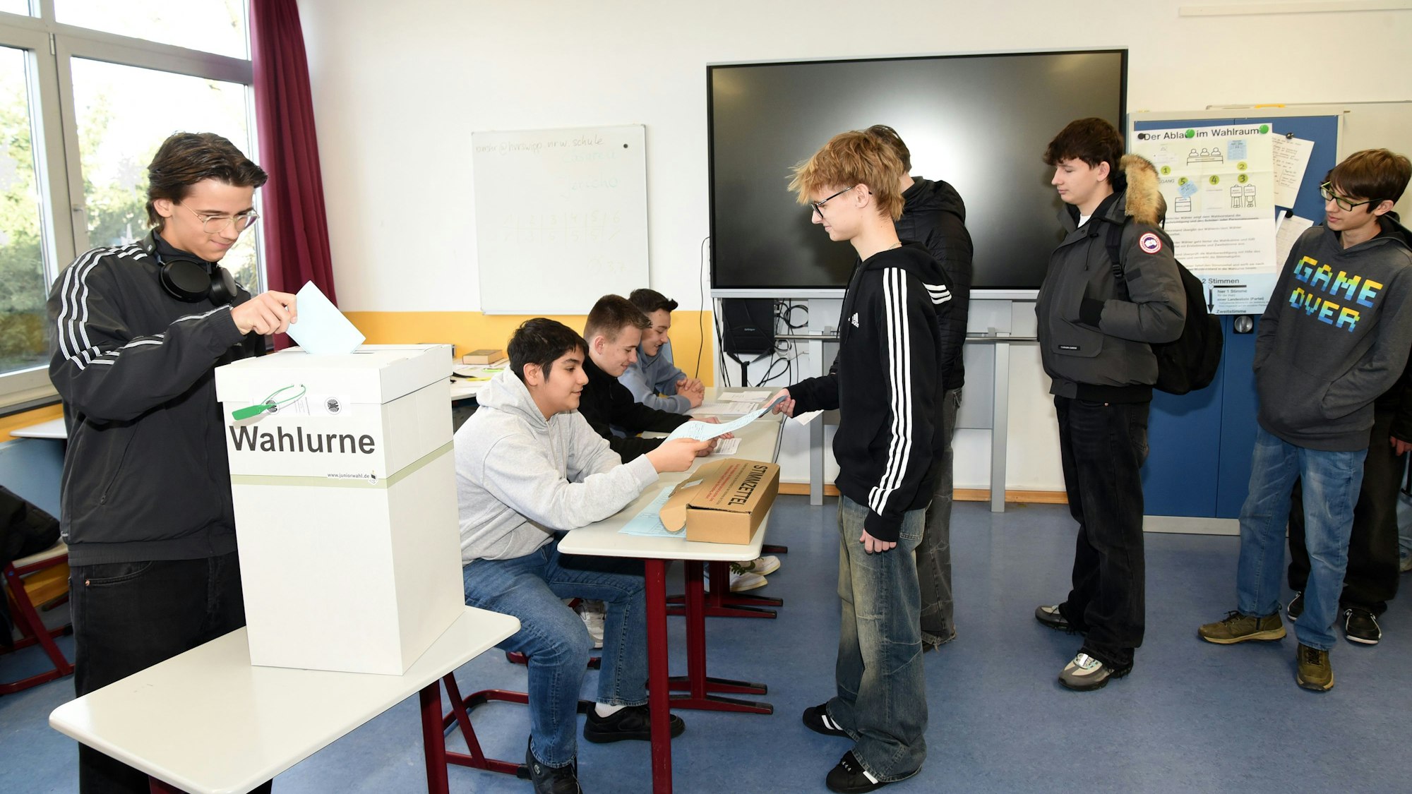 Mehrere Schüler stehen an einer Wahlurne in einem Klassenzimmer an, in die sie ihre Wahlzettel schmeißen.