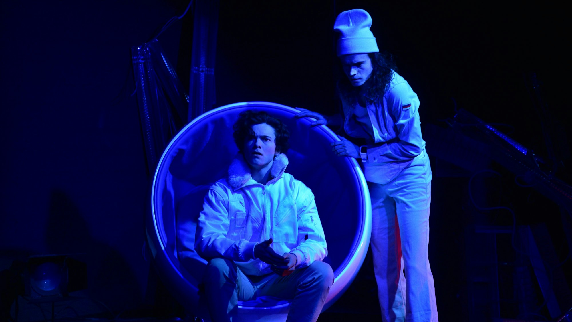 Zwei Schauspieler auf der Bühne, das Licht ist blau eingefärbt. Einer sitzt in einem Sessel.