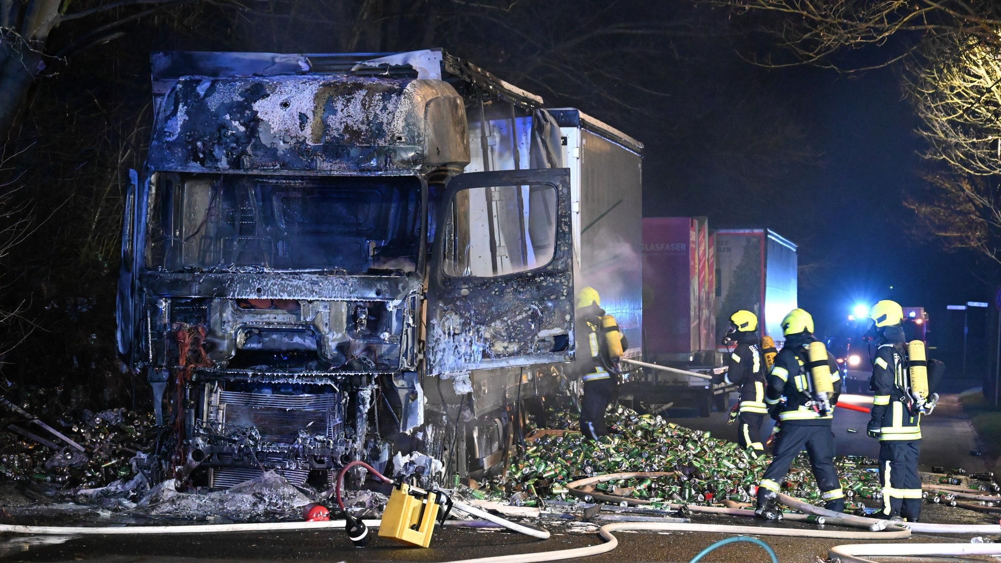 Ein Lkw steht mit völlig ausgebranntem Führerhaus am Straßenrand. Von rechts führen Feuerwehrleute letzte Löscharbeiten durch.