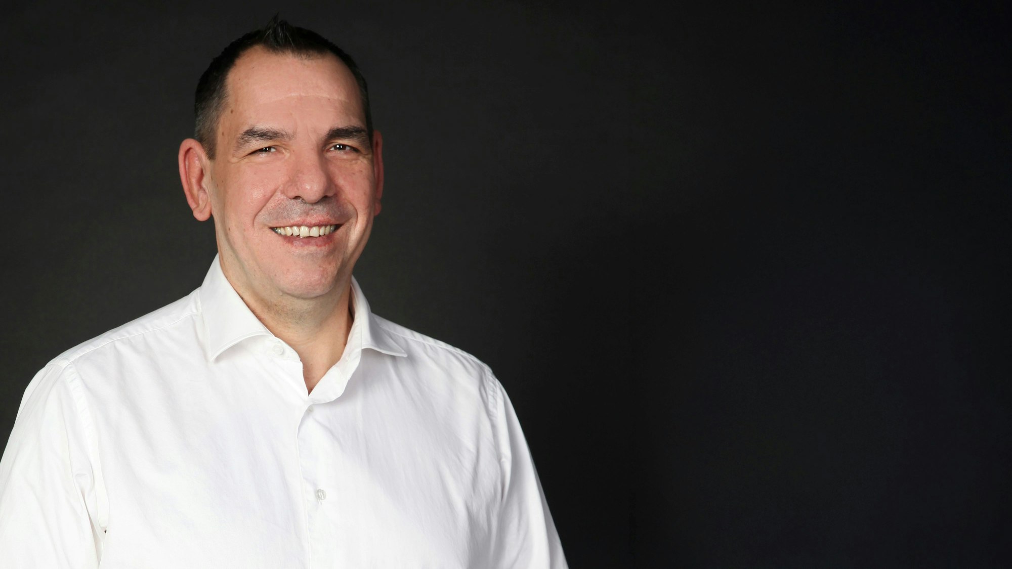 Das Bild zeigt Thomas Wiza. Er trägt ein weißes Hemd und steht vor einer schwarzen Wand.