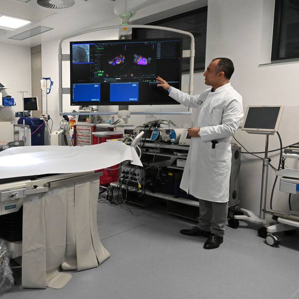 Dr. Erol Saygili, der Chefarzt der Kardiologie am Kreiskrankenhaus in Mechernich, trägt einen weißen Kittel und steht in einem Herzkatheter-Labor, das mit vielen technischen Geräten und einem großen Bildschirm ausgestattet ist.