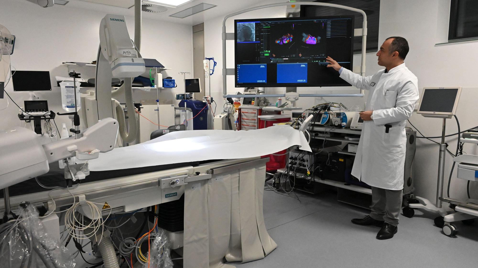 Dr. Erol Saygili, der Chefarzt der Kardiologie am Kreiskrankenhaus in Mechernich, trägt einen weißen Kittel und steht in einem Herzkatheter-Labor, das mit vielen technischen Geräten und einem großen Bildschirm ausgestattet ist.