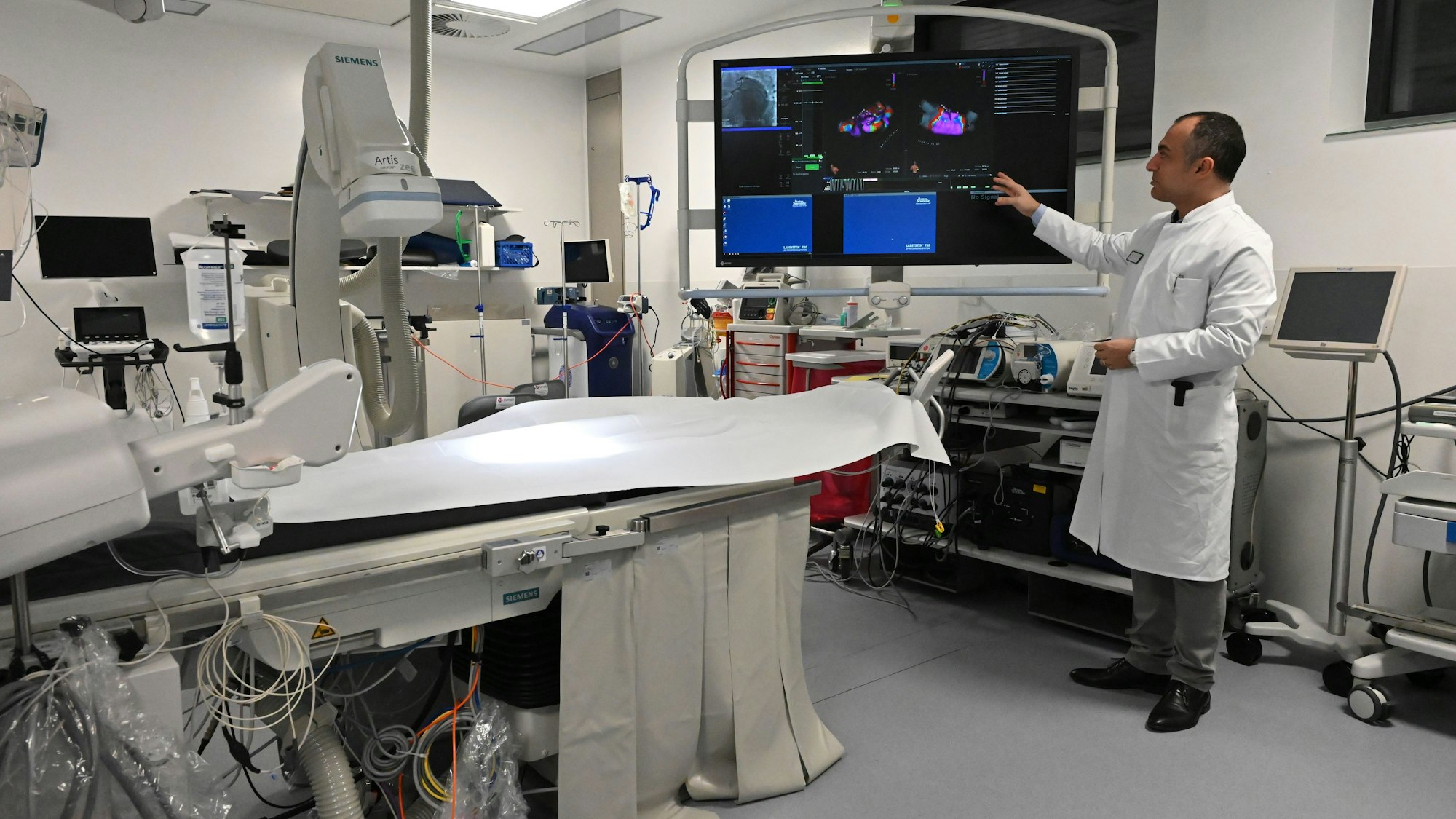 Dr. Erol Saygili, der Chefarzt der Kardiologie am Kreiskrankenhaus in Mechernich, trägt einen weißen Kittel und steht in einem Herzkatheter-Labor, das mit vielen technischen Geräten und einem großen Bildschirm ausgestattet ist.