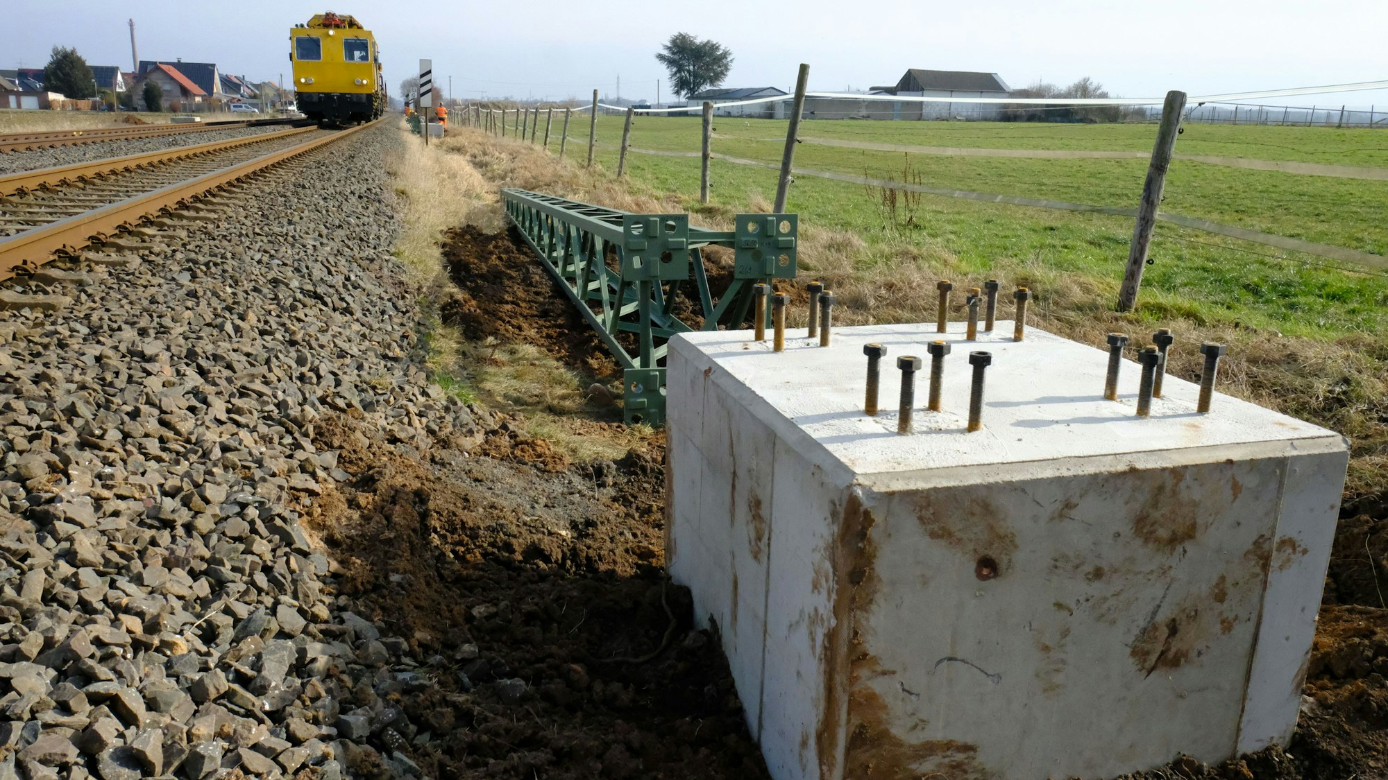 Neben einer Bahnstrecke befindet sich ein Betonfundament. Dahinter liegt ein Oberleitungsmast aus grün lackiertem Metall.