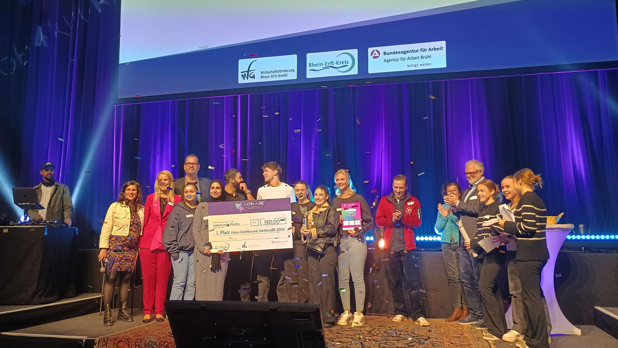 Die Schülerinnen und Schüler stehen mit der Jury auf der Bühne und halten ihren Scheck, während Konfetti herunterrieselt.