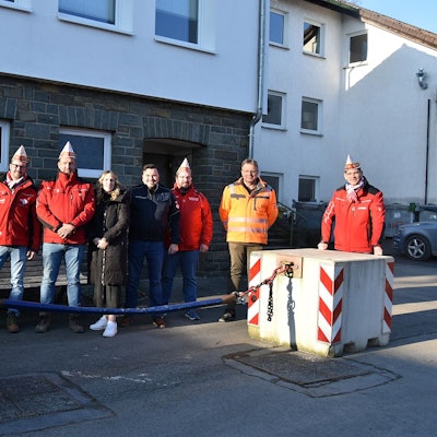 Mehrere Vereinsmitglieder eines Karnevalsvereins stehen auf der Straße neben einer Antiterrorsperre mit Sicherheitskette zwischen zwei Betonblöcken.