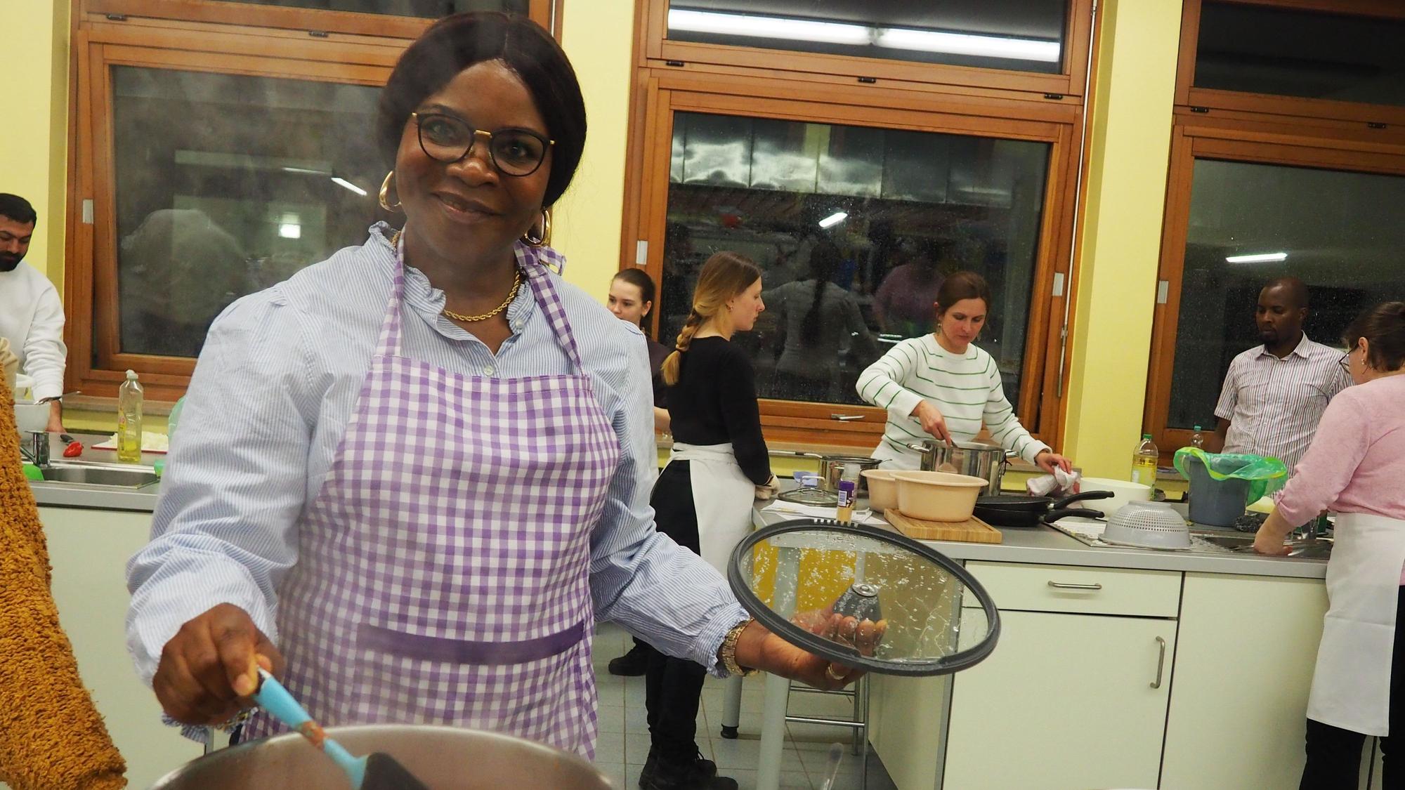Ikechi Alaike rührt in einem Topf, im Hintergrund sind weitere Menschen zu sehen, die kochen.