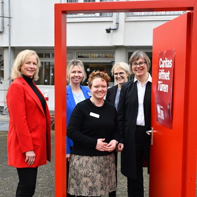 Das Foto zeigt das Führungsteam der Caritas vor einer geöffneten symbolischen roiten Tür.