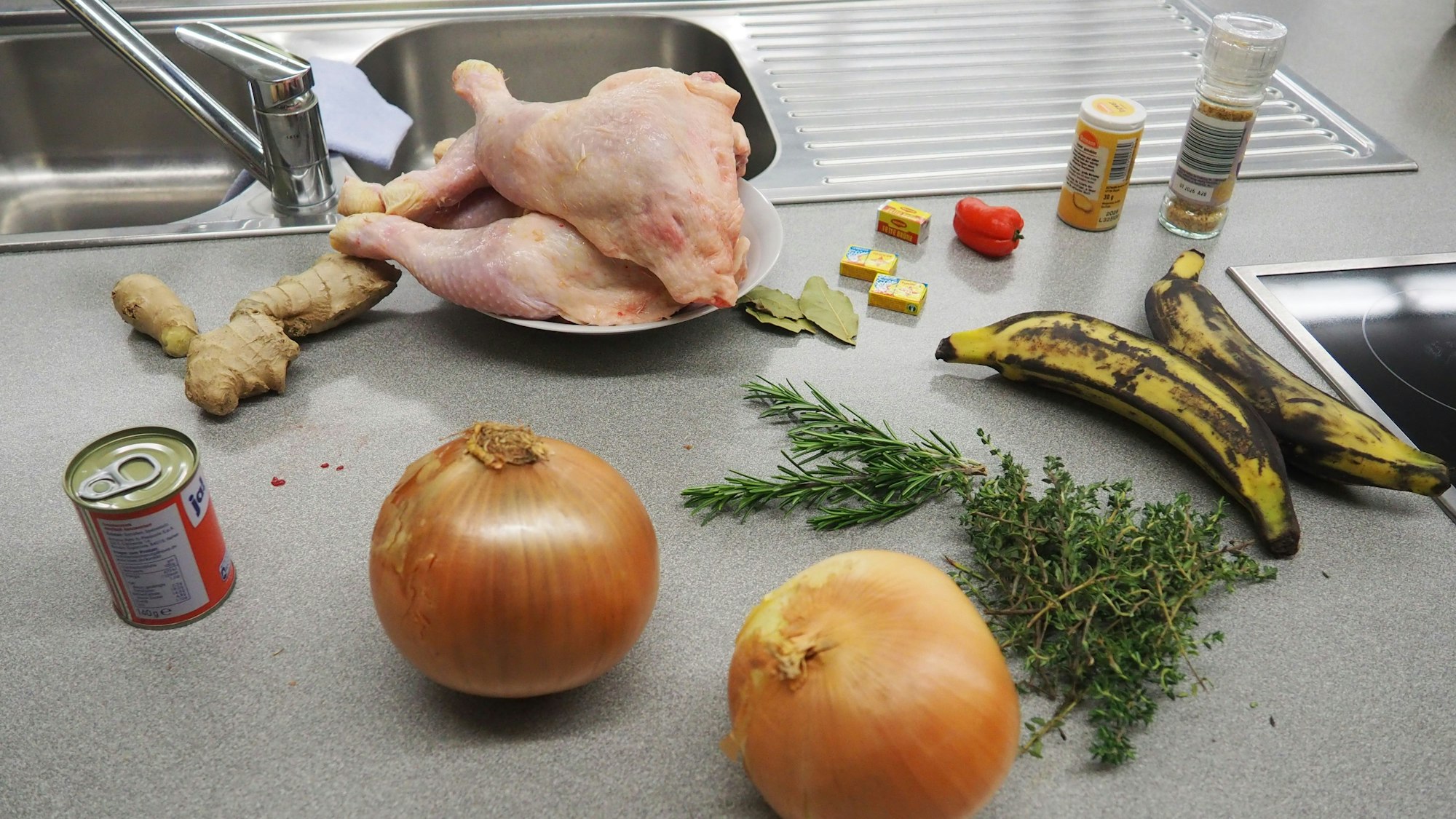 Zwiebeln, Gewürze, Kräuter und Hühnchenteile liegen auf der Arbeitsplatte einer Küche.