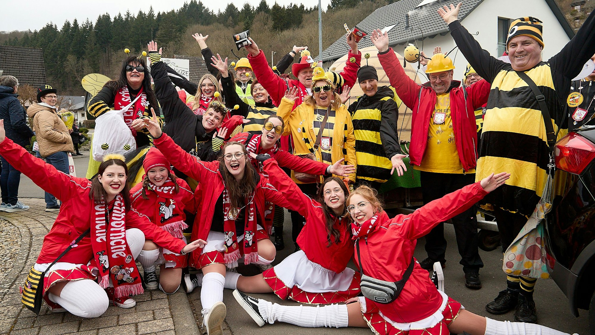 Jecken in Bienenkostümen stehen nebeneinander und reißen die Arme hoch. Davor sind Mädchen in Gardeuniformen.