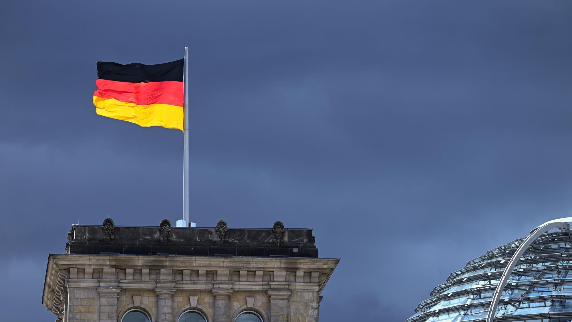 ARCHIV - 03.08.2023, Berlin: Eine der Flaggen auf dem Reichstag leuchtet bei starkem Wind wehend vor der Kulisse von dunklen Regenwolken.