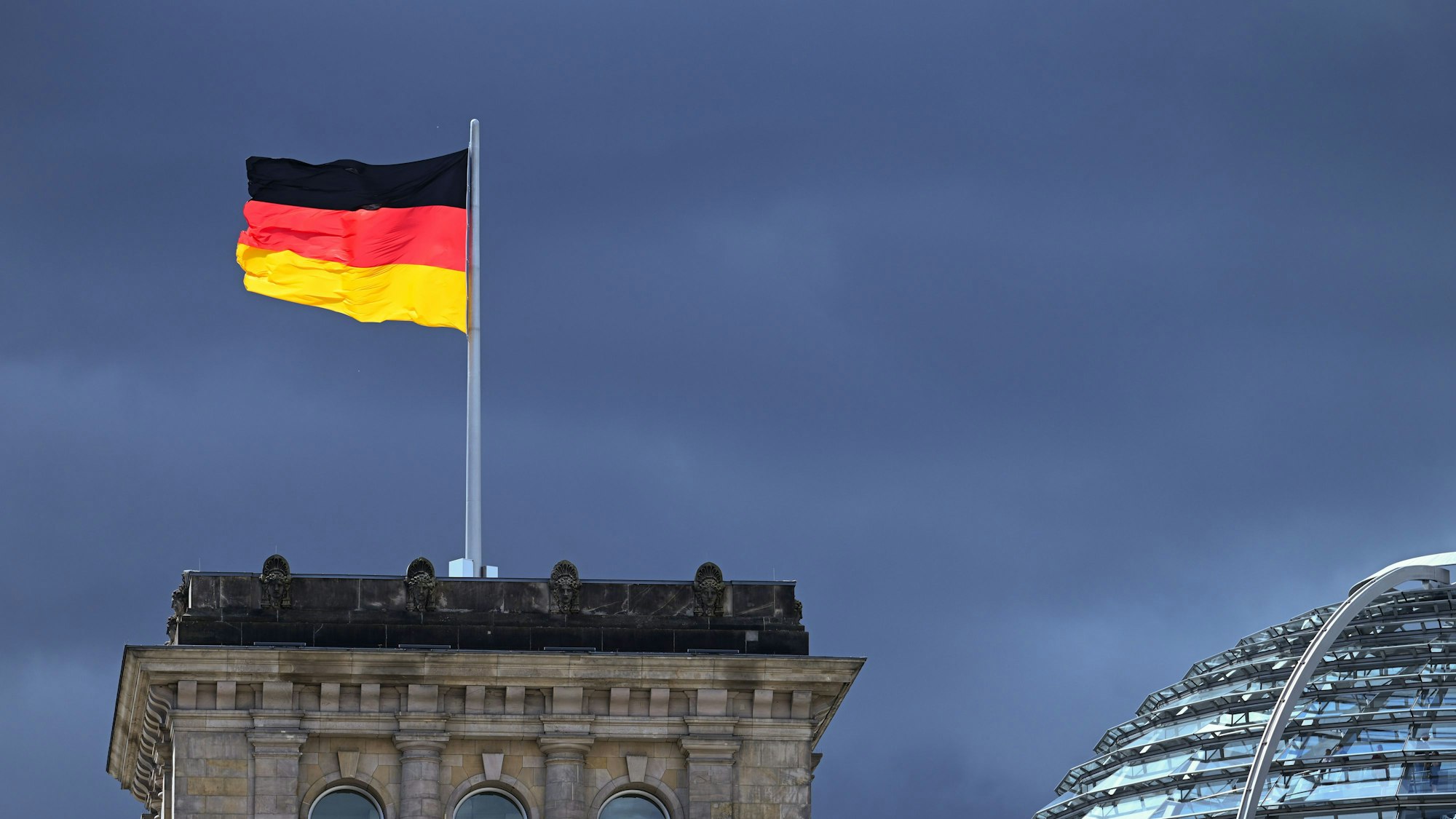 ARCHIV - 03.08.2023, Berlin: Eine der Flaggen auf dem Reichstag leuchtet bei starkem Wind wehend vor der Kulisse von dunklen Regenwolken.