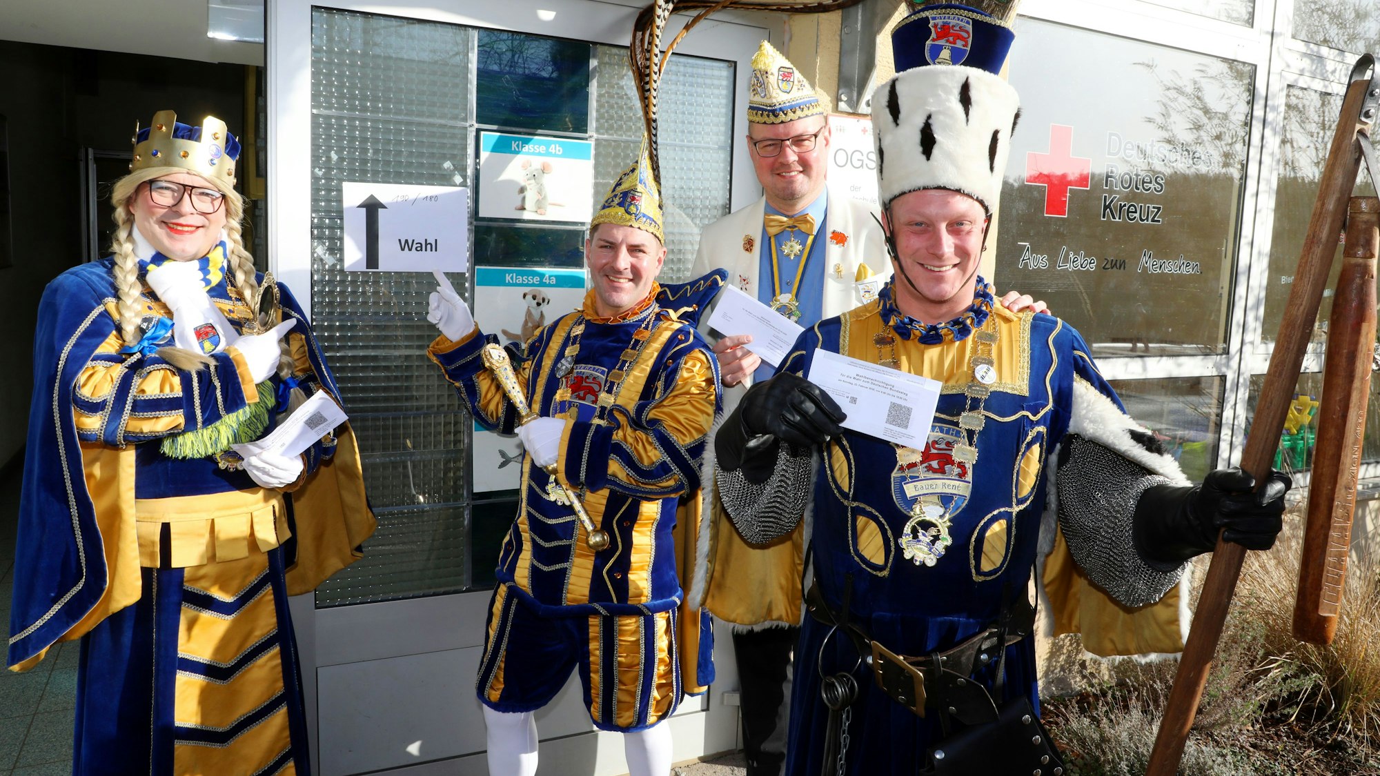 Jungfrau Timona (Timo Boxberg), Prinz Andreas I. (Lüdenbach), Prinzenführer Niklas Habers und Bauer René (Doppstadt) vom Overather Dreigestirn stehen vor einem Wahllokal in der Grundschule von Vilkerath.