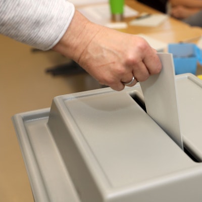 Das Foto zeigt einen Wahlzettel, den ein Wähler in eine Wahlurne gibt