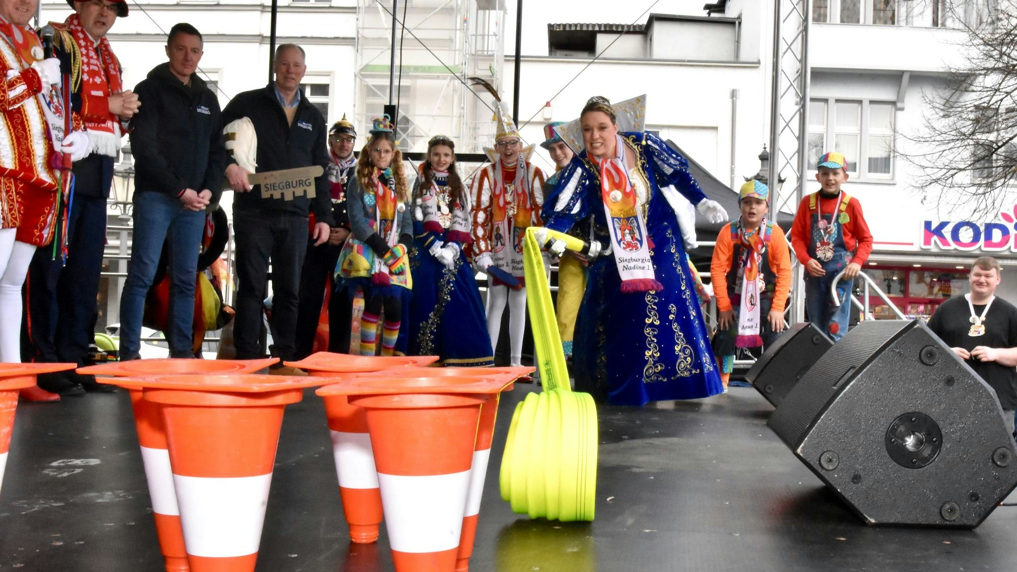 Eine Karnevalsprinzessin muss mit einem Feuerwehrschlauch Verkehrsleitkegel abräumen.