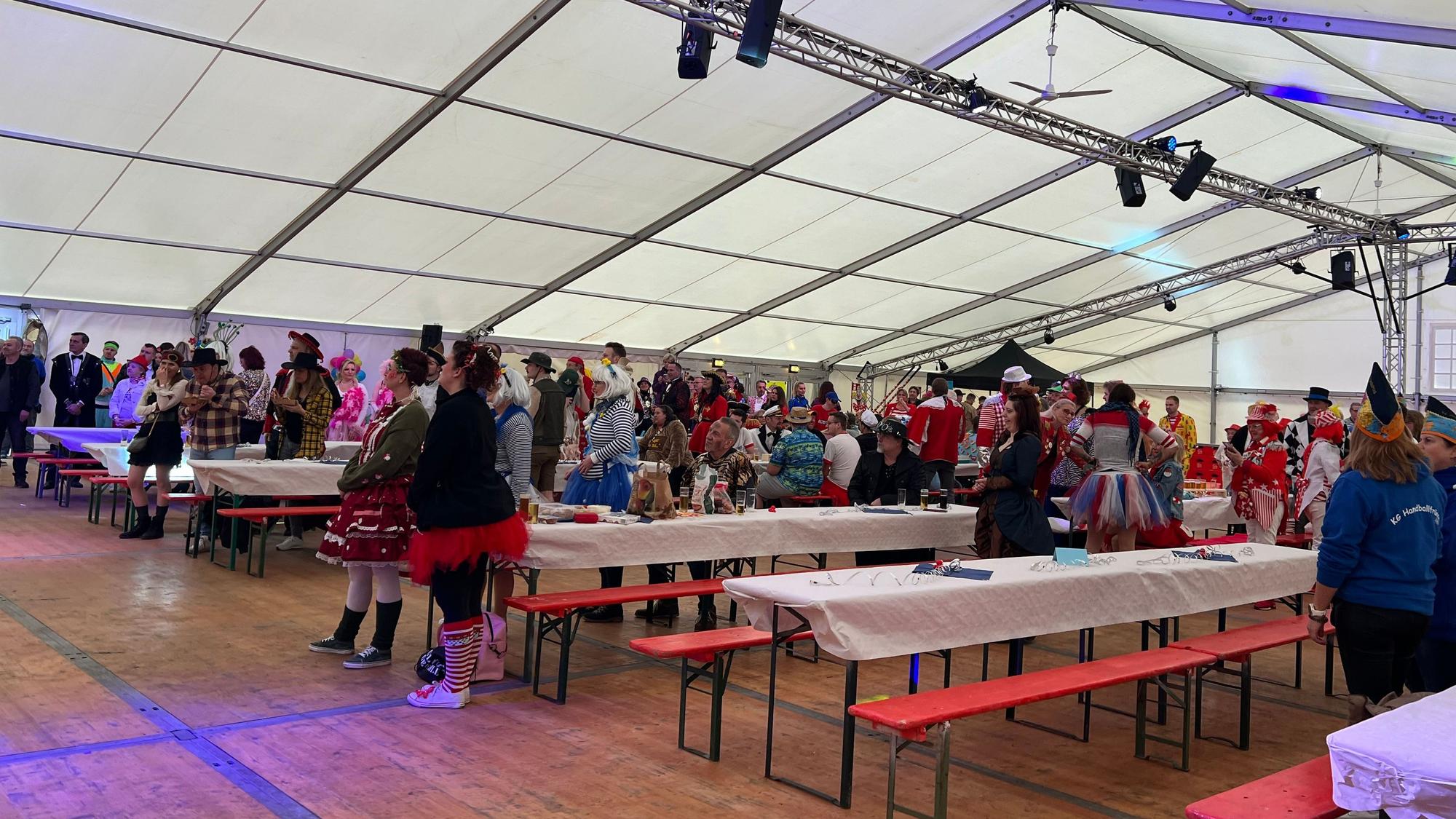 Kostümierte Karnevalisten stehen an halbleeren Biertischen in einem Zelt.