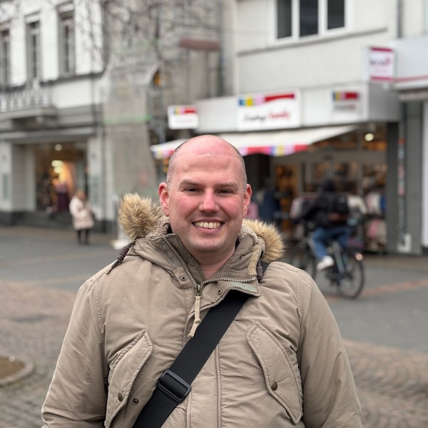 Ein Mann in einer Jacke steht in einer Fußgängerzone und zeigt seine Zähne beim Lächeln. Im Hintergrund befinden sich Passanten, die unscharf gestellt sind.