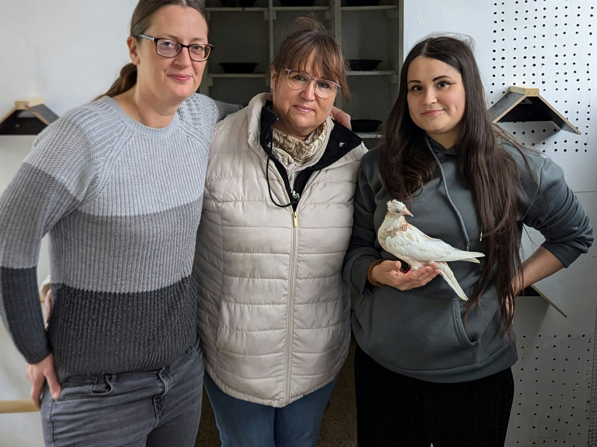 Drei Frauen stehen nebeneinander in einem Taubenhaus. Die ganz rechts hat eine braun-weiße Taube auf der Hand.