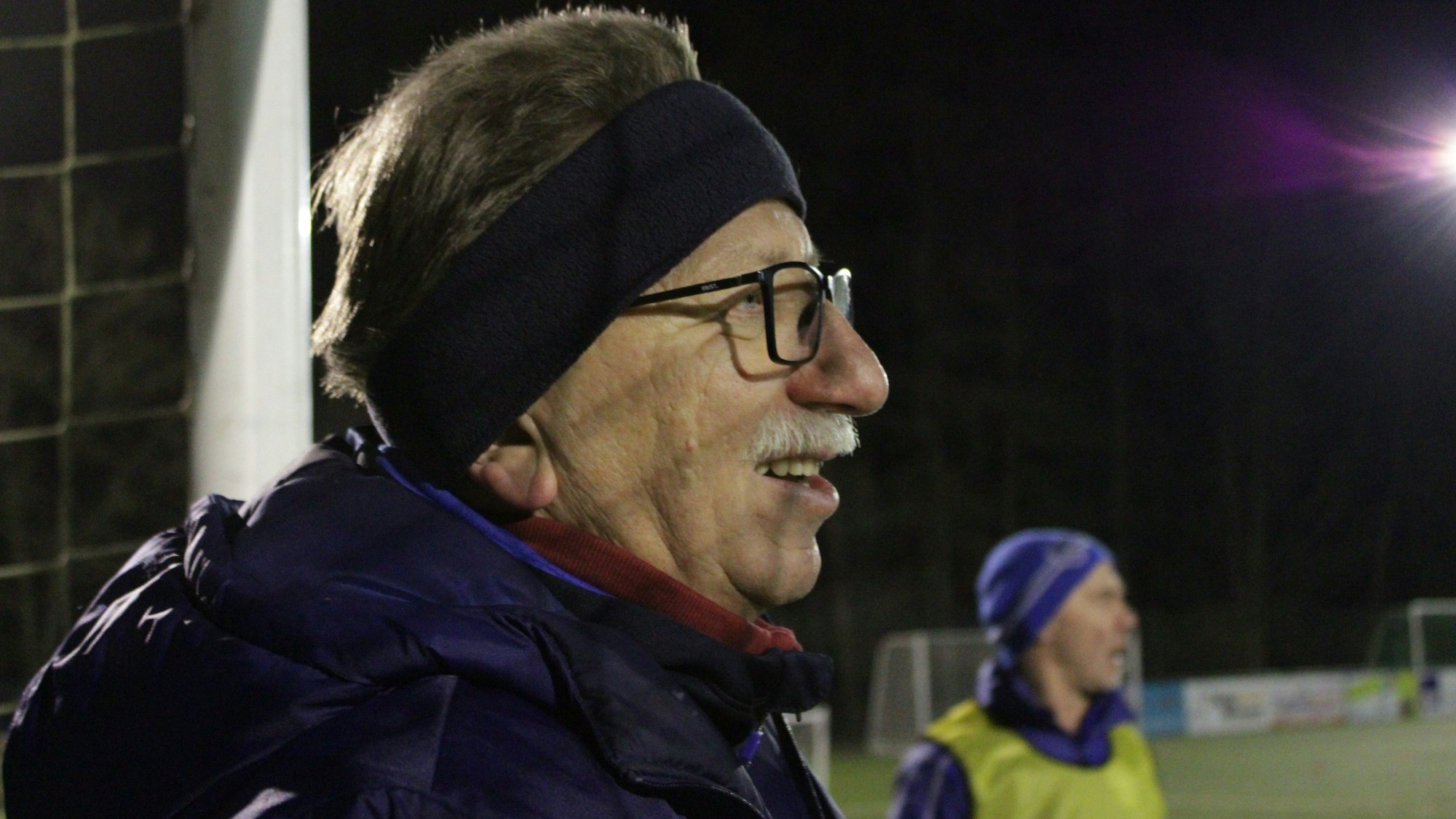 Ein älterer Mann mit Stirnband und Brille steht am Rand eines Fußballfeldes.