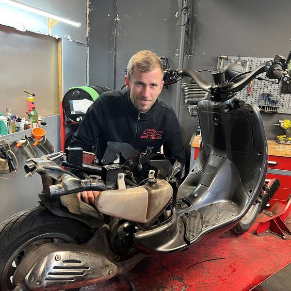 Ein blonder junger Mann steht in einer Werkstatt hinter einem Motorroller, den er gerade repariert.