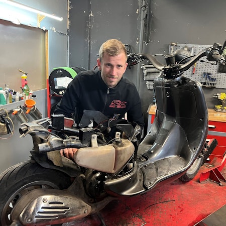 Ein blonder junger Mann steht in einer Werkstatt hinter einem Motorroller, den er gerade repariert.