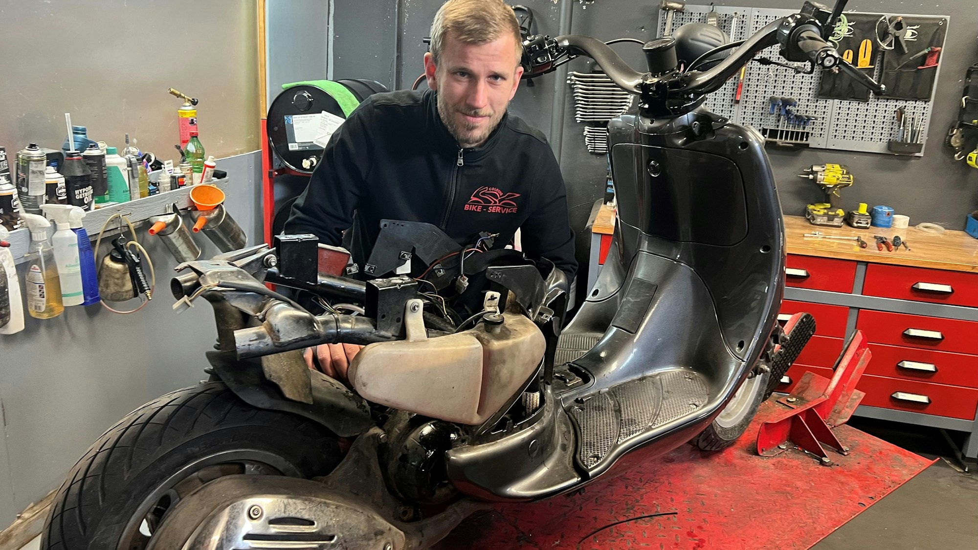 Ein blonder junger Mann steht in einer Werkstatt hinter einem Motorroller, den er gerade repariert.