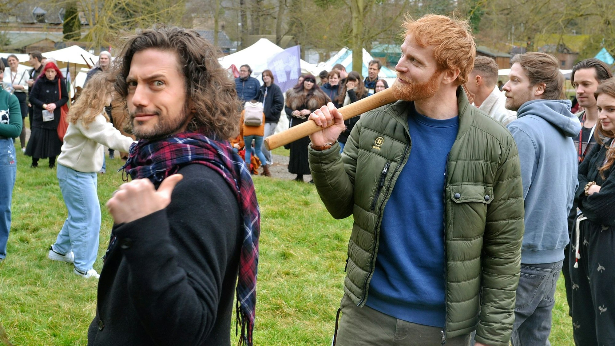 Die Schauspieler Jackson Rathbone in schwarzer Jacke und Magnus Bruun in grüner Jacke stehen auf einer Wieser der Burg Satzvey. Im Hintergrund sind zahlreiche Fans zu sehen.