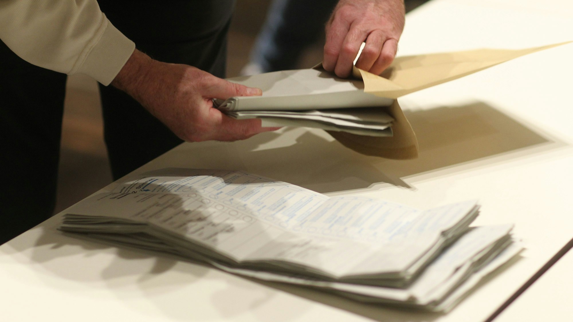 Ein Stapel Stimmzettel liegt auf einem Tisch, ein Mann steckt Stimmzettel in ein Kuvert.