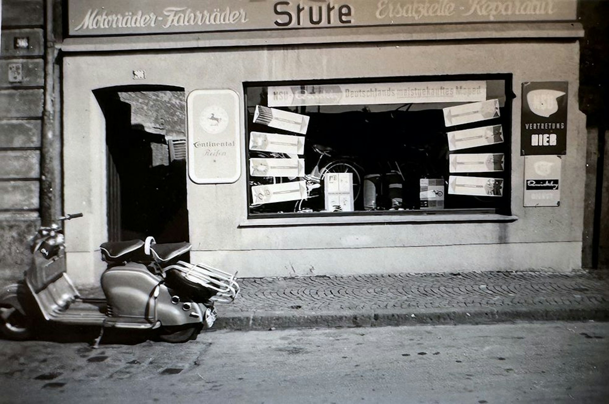 Blick auf einen kleinen Laden mit Schaufenster, davor parkt eine Vespa.