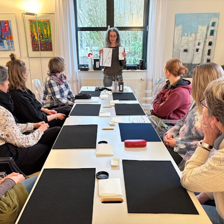Eine Frau mit Brille steht vor sieben Frauen, die an einem Tisch sitzen und ihr zugewandt sind. Sie hält zwei Bilder in die Luft.