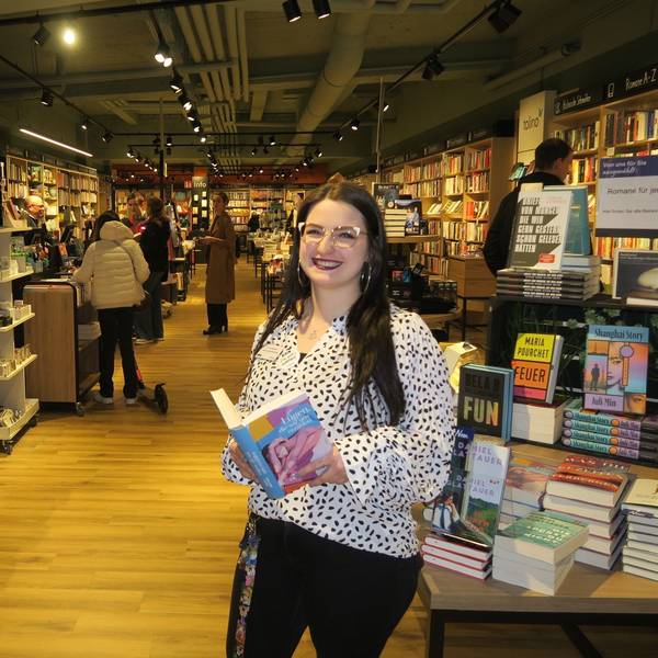 Eine junge Frau mit langem schwarzem Haar steht an einem Büchertisch mit einem aufgeschlagenen Buch in der Hand.