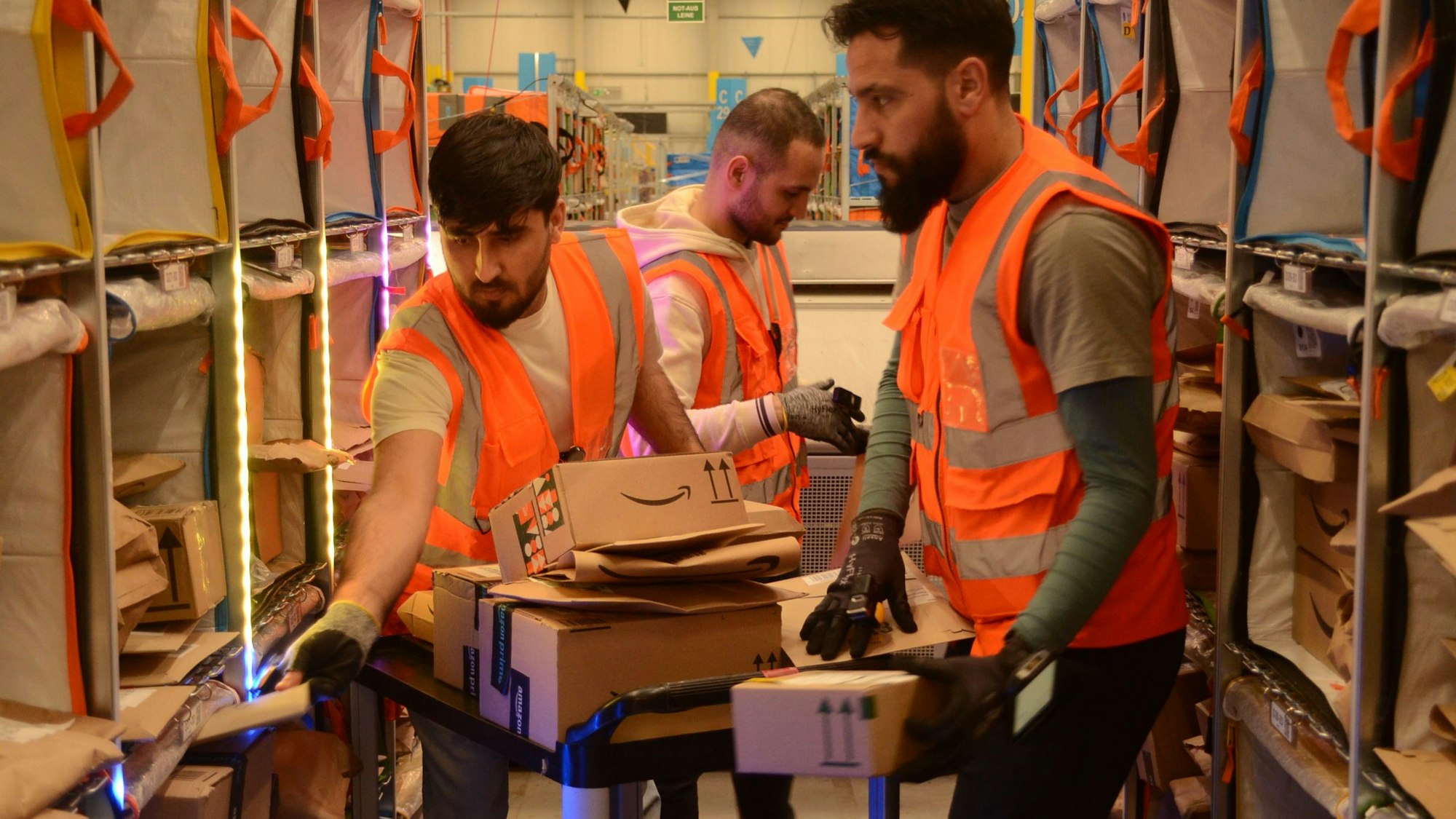 Drei Männer stehen im Gang zwischen Regalen und sortieren Pakete und Päckchen in die einzelnen Fächer.