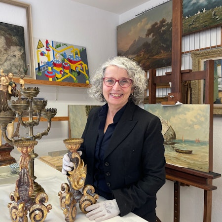 Eine Frau mit schwarzer Jacke und grauem Haar steht an einem Tisch. Sie hält einen antiken Kerzenleuchter in den behandschuhten Händen.