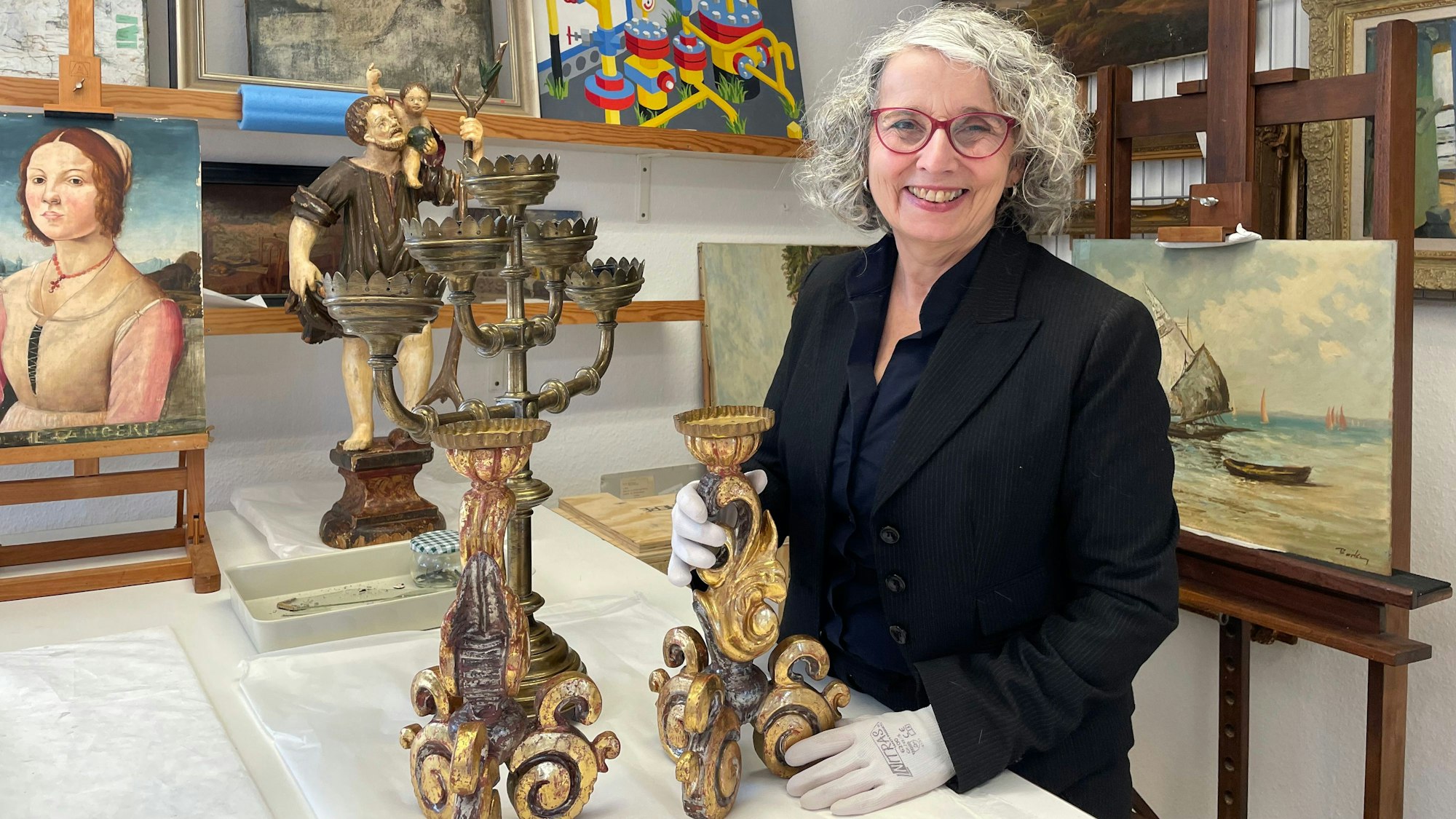 Eine Frau mit schwarzer Jacke und grauem Haar steht an einem Tisch. Sie hält einen antiken Kerzenleuchter in den behandschuhten Händen.