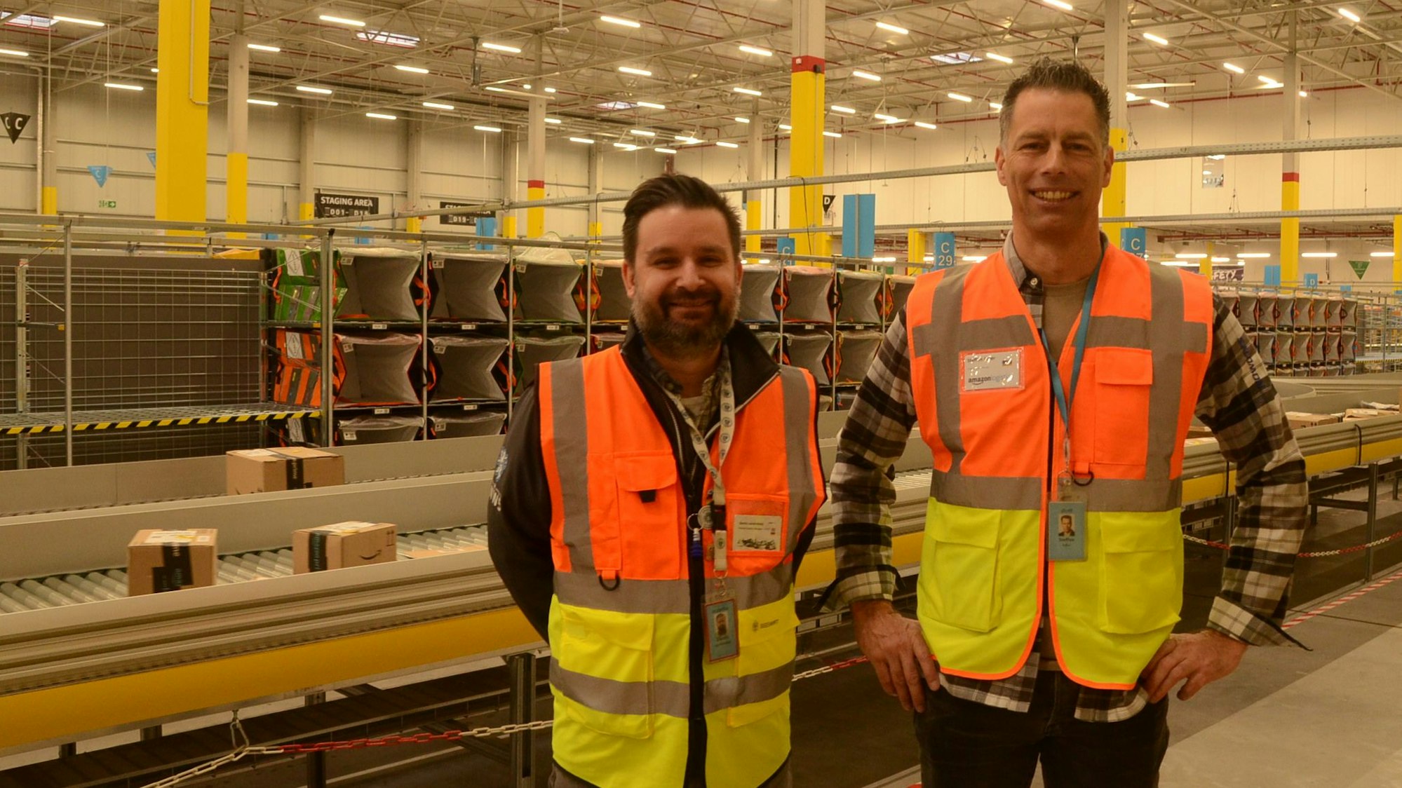 Zwei Männer in orange-gelben Warnwesten stehen vor einem Förderband, auf dem Pakete transportiert werden.