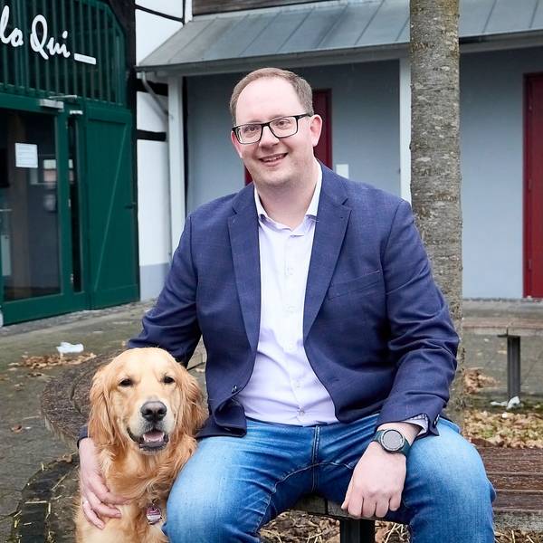 Sebastian Schubert sitzt auf einer Bank vor einem Baum. Mit einer Hand streichelt er einen Golden Retriever.