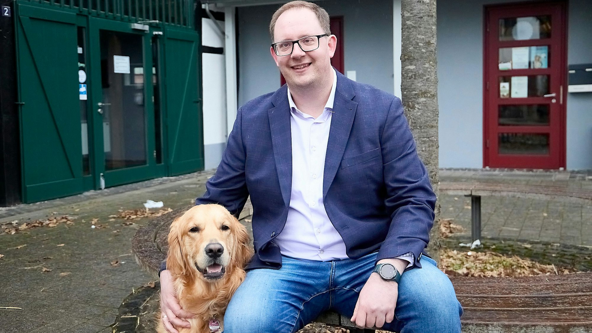 Sebastian Schubert sitzt auf einer Bank vor einem Baum. Mit einer Hand streichelt er einen Golden Retriever.