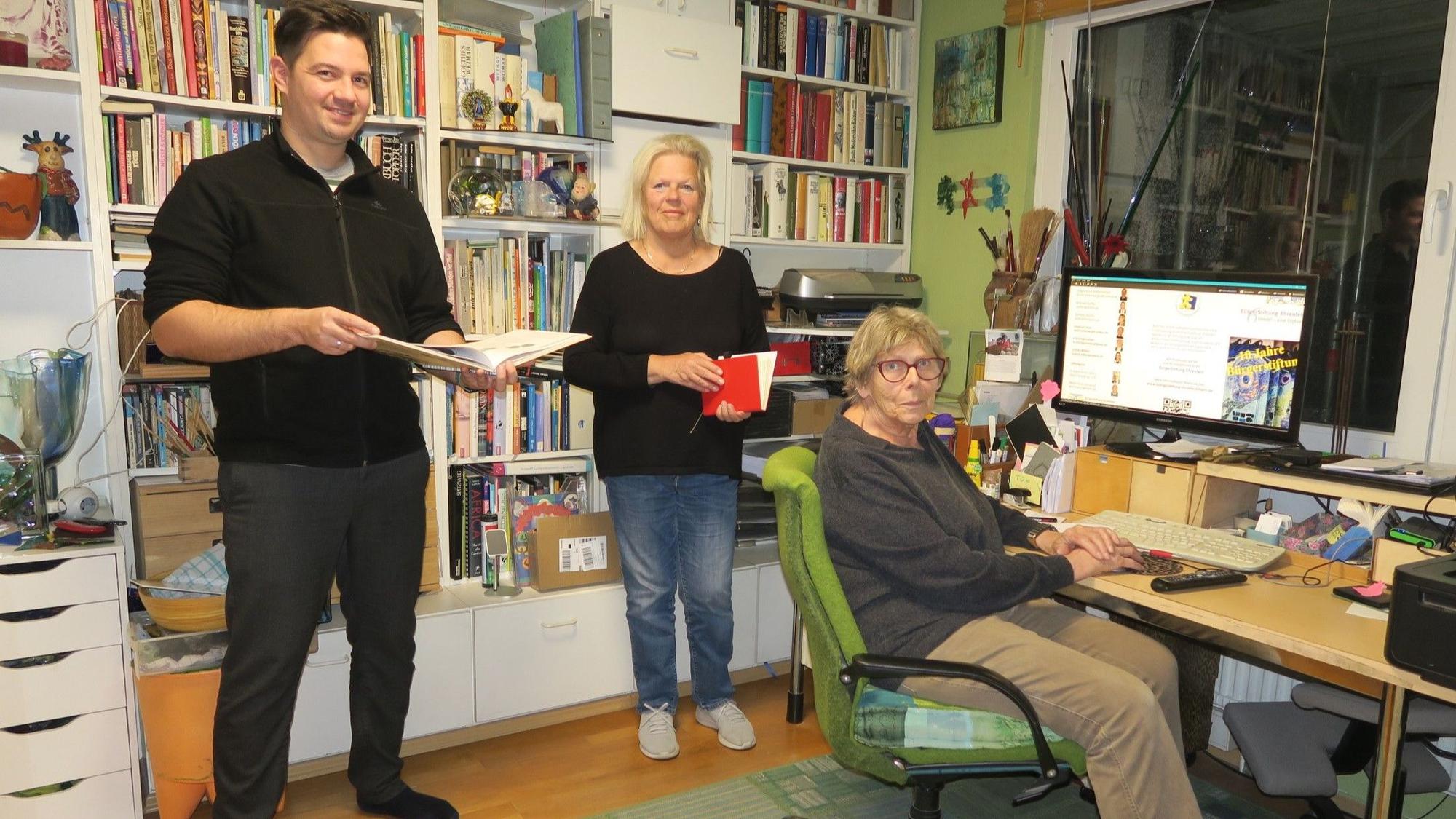 Ein Mann und eine Frau stehen in einem Arbeitszimmer, eine ältere Frau sitzt an einem Schreibtisch mit großem Monitor.