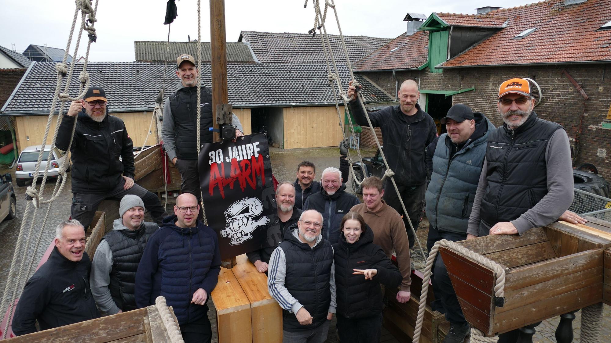 13 Männer und ein Mädchen in dunklen Klamotten stehen auf einem Holzschiff.
